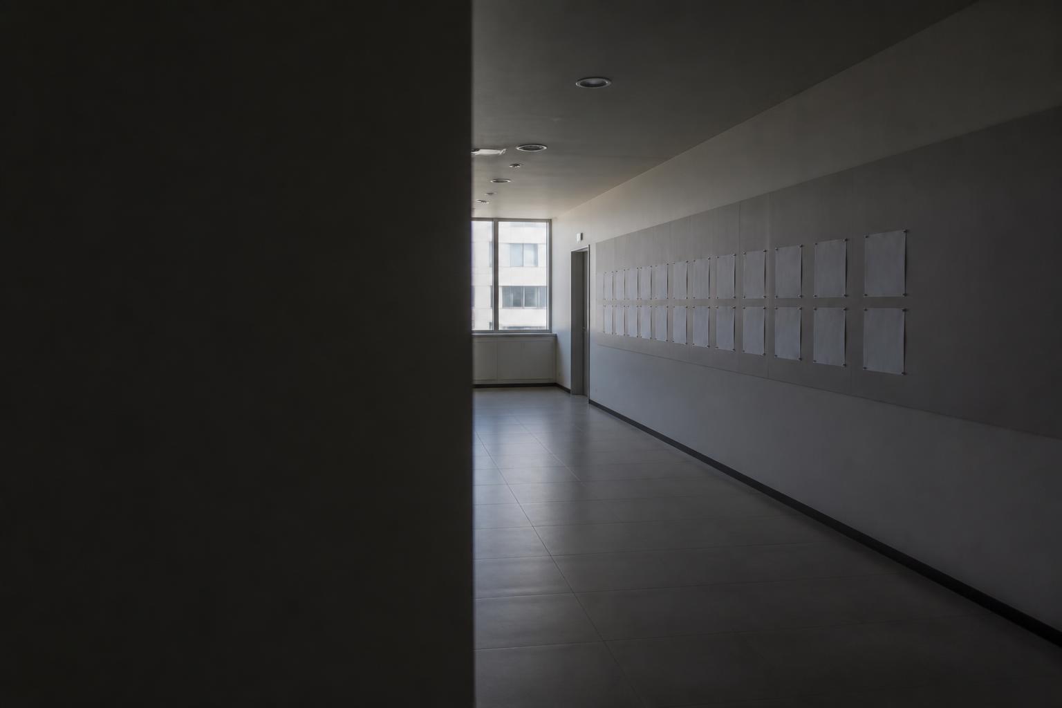끝 창에서 볕이 드는 복도. 오른쪽 벽에 빈 종이가 줄지어 붙어 있고 왼쪽은 그늘진 벽이다